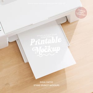 Puede incluir: Una impresora blanca con una pila de papel blanco. La hoja superior tiene las palabras "Printable Mockup" en una fuente estilizada. La imagen también incluye el texto "STYLED MOCKUP SCENE" y "STYKKE STUDIO". El fondo es una superficie de madera clara.