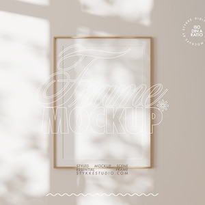 Puede incluir: Una maqueta de marco rectangular dorado con el texto "Frame MOCKUP" en una fuente blanca. El marco está colgado en una pared beige con sombras. El texto "STYLED MOCKUP SCENE ESSENTIAL FRAME STYKKESTUDIO.COM" está en la parte inferior.