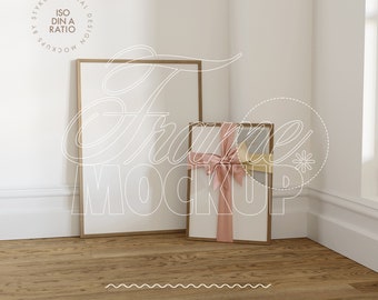 May include: Two wooden picture frames, one larger than the other, are leaning against a white wall. The smaller frame is decorated with a pink ribbon. The frames are on a wooden floor.