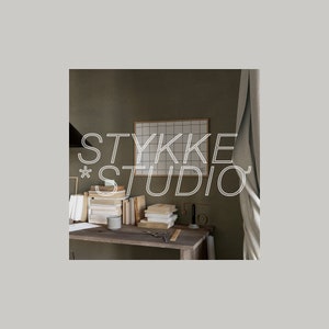 May include: A close-up of a desk with a stack of books, a white gridded board, and the text "STYKKE STUDIO" in white lettering. The desk is made of wood and has a light brown finish. The background is a light brown wall.