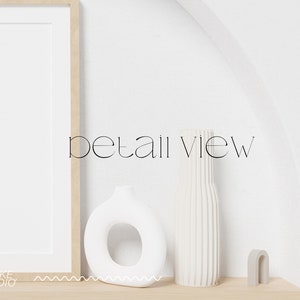 May include: A white ceramic vase with a ribbed texture, a white ceramic ring-shaped vase, and a gray arch-shaped sculpture on a light wood shelf. The text "detail view" is written in black on a white wall.