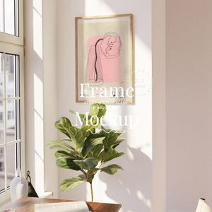 May include: A framed art print with a pink and black floral design. The artwork is in a gold frame and is on a white wall. The text "Frame" and "Mockup" are visible. A green plant is in front of the artwork. The scene is lit by natural light.