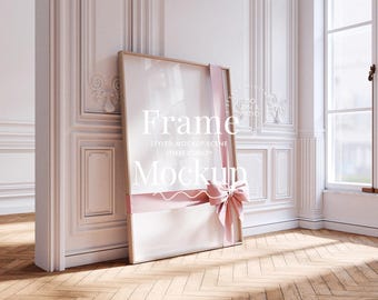 May include: A white frame mockup with a pink ribbon and bow, leaning against a white wall with ornate moulding. The frame is in a room with a wood floor and a window.