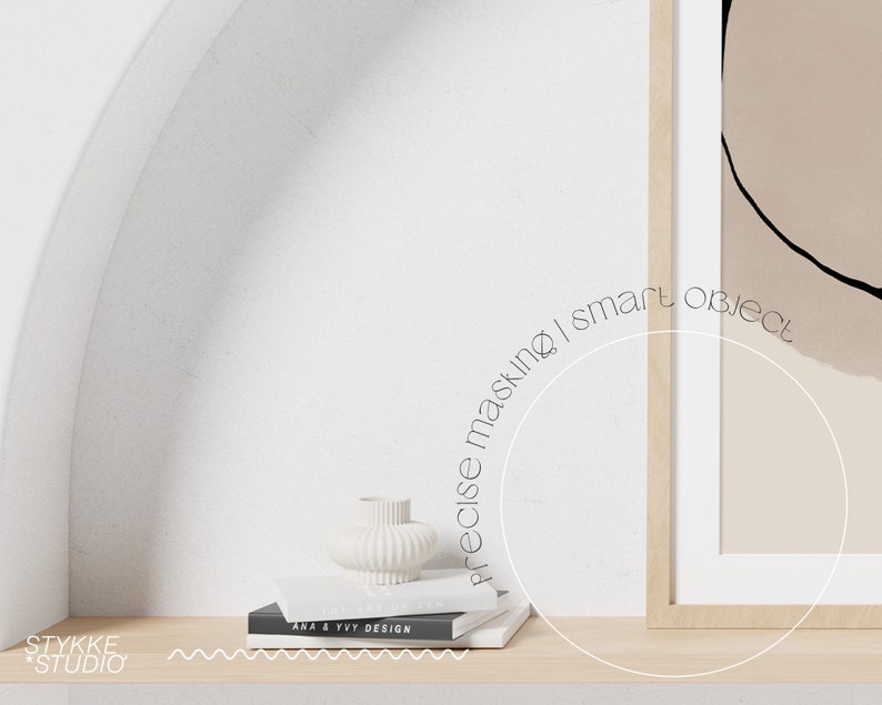 May include: A white ceramic vase with a ribbed texture sits on a stack of books with the text "ANA & VIV DESIGN" on the top book. The books are on a light wood shelf in front of a white wall with an archway. A framed abstract art print with a beige background and black lines hangs on the wall.
