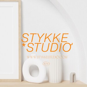 May include: A white framed picture with a white background. The text "STYKKE * STUDIO" in orange is below the frame. The text "WWW.STYKKE STUDIO.COM" is below the orange text. A white vase with vertical lines is on a wooden shelf. A white ring-shaped object is to the left of the vase. A small gray arch-shaped object is to the right of the vase.