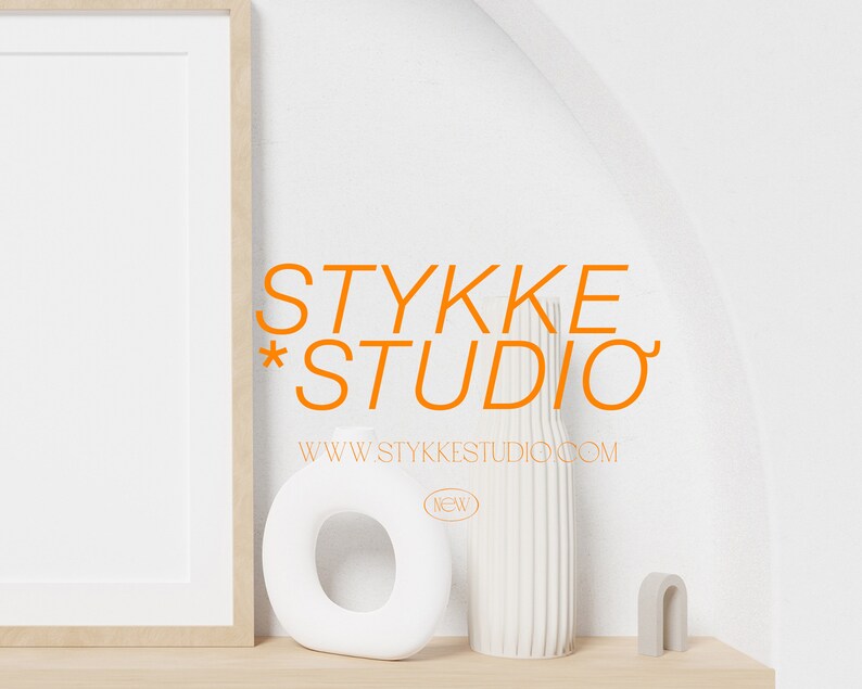 May include: A white framed picture with a white background. The text "STYKKE * STUDIO" in orange is below the frame. The text "WWW.STYKKE STUDIO.COM" is below the orange text. A white vase with vertical lines is on a wooden shelf. A white ring-shaped object is to the left of the vase. A small gray arch-shaped object is to the right of the vase.