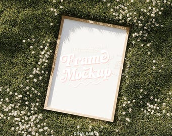 May include: A rectangular frame mockup with a white interior, set against a backdrop of green grass and small white flowers. The frame has a light brown border. The text "Frame Mockup" is displayed in a pink, stylized font.