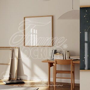 Pode incluir: Uma maquete de moldura de madeira quadrada com fundo branco, sentada em uma parede branca em uma sala com piso de madeira. Há uma mesa com uma cadeira e livros na mesa. Uma grande impressão emoldurada em preto e dourado com um design celestial está na parede à direita da maquete da moldura.