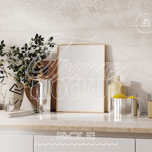 Könnte beinhalten: Eine stilvolle Mockup-Szene mit einem leeren Bilderrahmen mit Holzrahmen, einer Vase mit grünen Blättern, einem Glas Wasser, einem Holzbrett, einem Glaszylinder, einer Schüssel mit Zitronen und zwei Gläsern auf einer weißen Arbeitsplatte mit einer Betonrückwand. Der Text "Styled Mockup Scene Concrete Set No. 19 Stykkestudio.com" ist auf die Arbeitsplatte gedruckt.