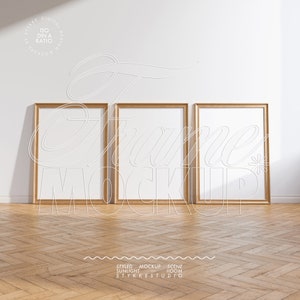 May include: Three gold-framed rectangular mockups are displayed against a white wall, set on a wooden parquet floor. The frames are empty, with the words "Frame Mockup" overlaid in a decorative font. The image includes the text "Styled Mockup Scene Sunlight Room STYKKESTUDIO".