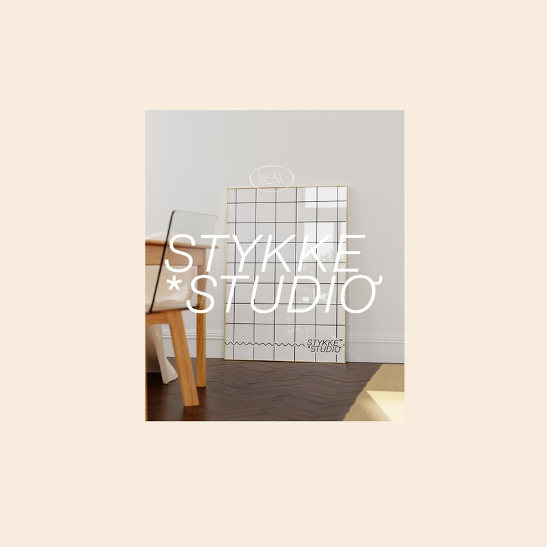 May include: A white gridded poster with a gold frame, hanging on a white wall. The poster has the text "STYKKE STUDIO" in white letters. The poster is hanging in a room with a wooden floor and a wooden chair.