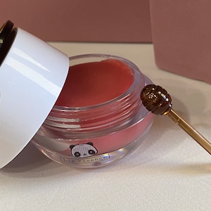 Könnte beinhalten: Ein transparentes Glas mit einem rosa Lippenbalsam darin. Das Glas hat ein schwarz-weißes Pandadesign mit dem Text "lip essence". Ein kleiner goldener und brauner Honiglöffel liegt neben dem Glas.