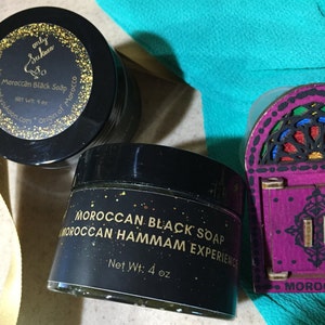 Puede incluir: Dos frascos negros de jab&oacute;n negro marroqu&iacute; con letras doradas. Los frascos est&aacute;n etiquetados como "Jab&oacute;n negro marroqu&iacute;" y "Experiencia Hammam marroqu&iacute;". Los frascos est&aacute;n sobre una mesa con una puerta de madera decorativa con una ventana de vitrales.