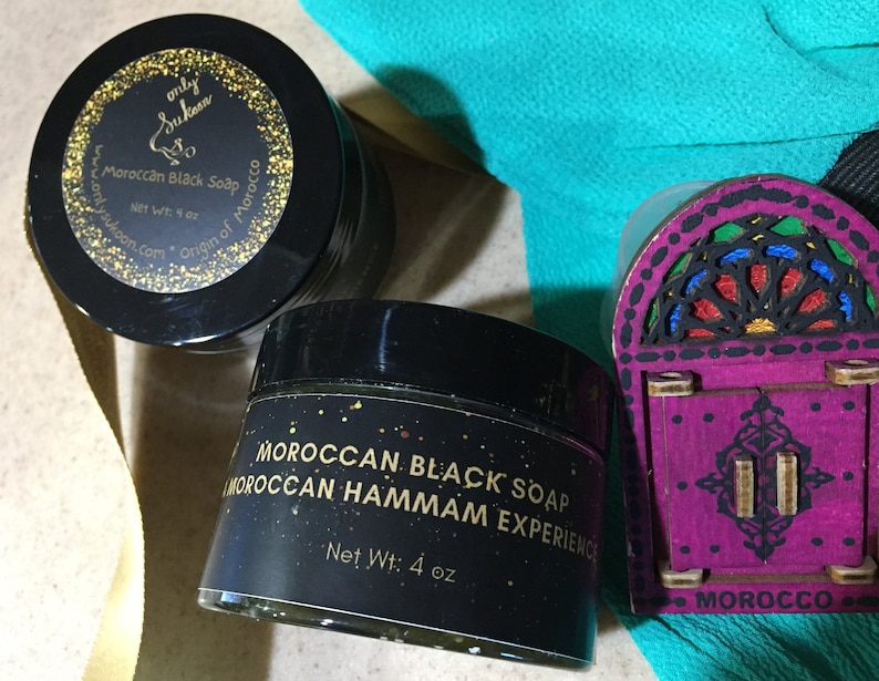 Puede incluir: Dos frascos negros de jab&oacute;n negro marroqu&iacute; con letras doradas. Los frascos est&aacute;n etiquetados como "Jab&oacute;n negro marroqu&iacute;" y "Experiencia Hammam marroqu&iacute;". Los frascos est&aacute;n sobre una mesa con una puerta de madera decorativa con una ventana de vitrales.
