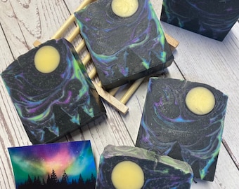 May include: Artisan soaps with a night sky design. The soaps are black with swirls of purple, blue, and green, and a yellow moon. A small card shows a colorful aurora borealis scene. The soaps are arranged on a wooden surface.