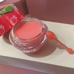 May include: A small, round, clear glass jar filled with a smooth, coral-colored lip balm. The jar has a red lid decorated with white strawberry outlines and a green accent. A small, translucent, coral-colored applicator wand is placed beside the jar.