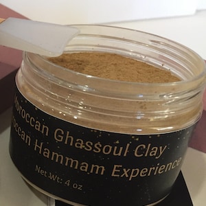 Puede incluir: Un tarro transparente con una etiqueta negra que dice "Moroccan Ghassoul Clay Hammam Experience" y "Net Wt. 4 oz". El tarro está lleno de un polvo marrón claro.