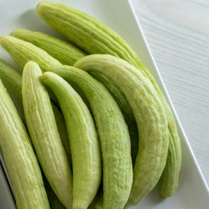 May include: A close-up of a bunch of light green cucumbers on a white plate. The cucumbers are arranged in a fan shape, with the tips of the cucumbers pointing towards the center of the plate.
