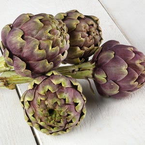 May include: Four fresh artichokes with purple and green hues rest on a white wooden surface. The artichokes are in various stages of bloom, showcasing their layered leaves and stems. The image highlights the natural textures and colors of the vegetables.