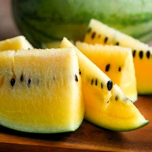 May include: Close-up of several slices of yellow watermelon with black seeds. The rind is green, and the flesh is a vibrant yellow. A whole watermelon is visible in the background.