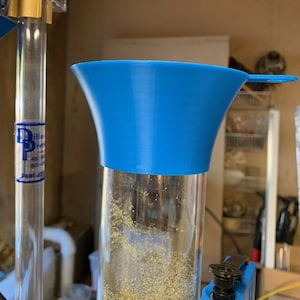 May include: A blue funnel sits atop a clear, cylindrical container filled with small, granular material. The funnel has a wide mouth and a small handle. The container is part of a larger apparatus, with additional components visible in the background.