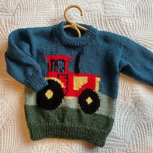 May include: A teal blue knitted sweater with a tractor design. The tractor is red, yellow, and black, set against a green and light blue background. The sweater is displayed on a wooden hanger.