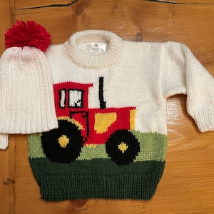 May include: A cream-colored knit sweater and matching hat set. The sweater features a red, yellow, and black tractor design on the front, with a green field at the bottom. The hat has a red pom-pom.