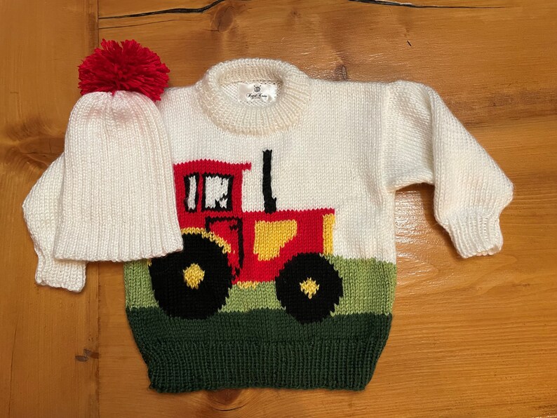 May include: A cream-colored knit sweater and matching hat set. The sweater features a red, yellow, and black tractor design on the front, with a green field at the bottom. The hat has a red pom-pom.