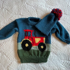 May include: A teal knitted sweater and matching hat set for a child. The sweater features a red tractor design with yellow and black accents. The hat has a red, navy, and teal pom-pom.
