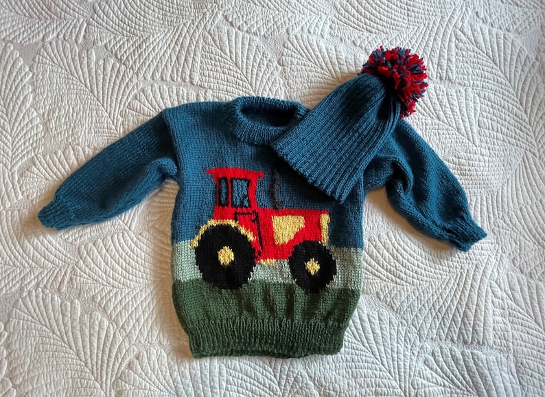 May include: A teal knitted sweater and matching hat set for a child. The sweater features a red tractor design with yellow and black accents. The hat has a red, navy, and teal pom-pom.