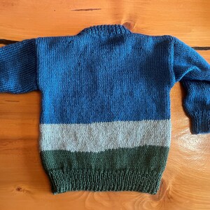 May include: A hand-knitted sweater in shades of blue, gray, and green. The sweater features a crew neck and long sleeves. The body of the sweater is divided into three horizontal stripes of color. The sweater is made of a soft, textured yarn.