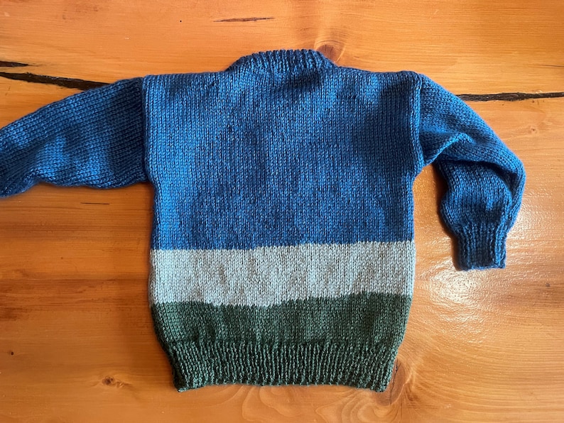 May include: A hand-knitted sweater in shades of blue, gray, and green. The sweater features a crew neck and long sleeves. The body of the sweater is divided into three horizontal stripes of color. The sweater is made of a soft, textured yarn.