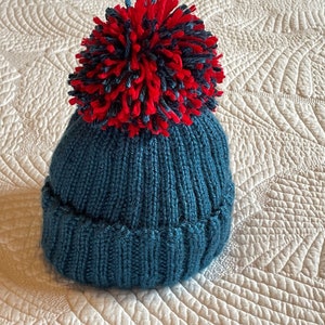 May include: A teal knitted beanie with a ribbed brim and a large, fluffy pom-pom in red and navy blue. The hat is made of a soft, textured yarn, perfect for cold weather. This winter hat is a cozy accessory.