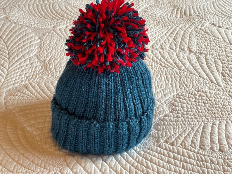 May include: A teal knitted beanie with a ribbed brim and a large, fluffy pom-pom in red and navy blue. The hat is made of a soft, textured yarn, perfect for cold weather. This winter hat is a cozy accessory.