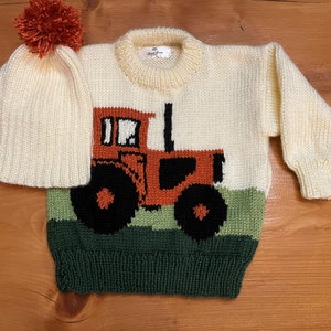 May include: Cream-colored knitted sweater and matching hat set. The sweater features a tractor design in orange, black, and green. The hat has a pom-pom on top. The set is designed for a baby or toddler.