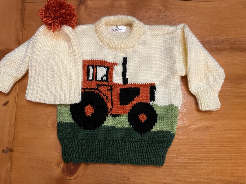 May include: Cream-colored knitted sweater and matching hat set. The sweater features a tractor design in orange, black, and green. The hat has a pom-pom on top. The set is designed for a baby or toddler.