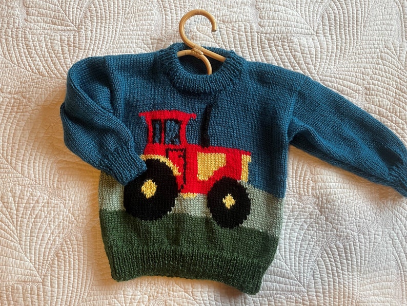 May include: A teal blue knitted sweater with a tractor design. The tractor is red, yellow, and black, set against a green and light blue background. The sweater is displayed on a wooden hanger.