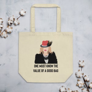 May include: A beige tote bag with a black and white illustration of a woman wearing a red hat and a black coat. The text "ONE MUST KNOW THE VALUE OF A GOOD BAG" is printed on the bag.