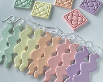 May include: A collection of wavy, matte-finish dangle earrings in pastel shades of mint green, pale yellow, peach, soft pink, and lavender, paired with matching square tiles featuring a raised five-petal flower design and silver-toned hooks.