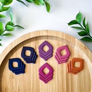 May include: Six layered geometric shapes in various shades of purple, pink, and orange on a wooden surface. The shapes are layered to create a three-dimensional effect.