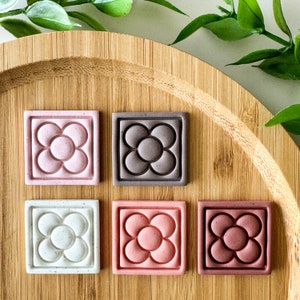 Puede incluir: Seis posavasos cuadrados en varios colores, incluyendo rosa, gris y blanco, cada uno con un diseño floral dentro de un marco cuadrado. Los posavasos están dispuestos sobre una bandeja de madera, con follaje verde en el fondo.