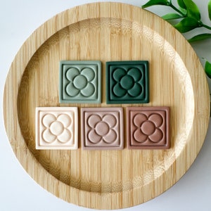 May include: Five square tiles featuring a raised five-petal flower design arranged in a row. The tiles are colored in shades of sage green, dark forest green, cream, dusty rose, and terracotta, each with a smooth, matte finish and border.