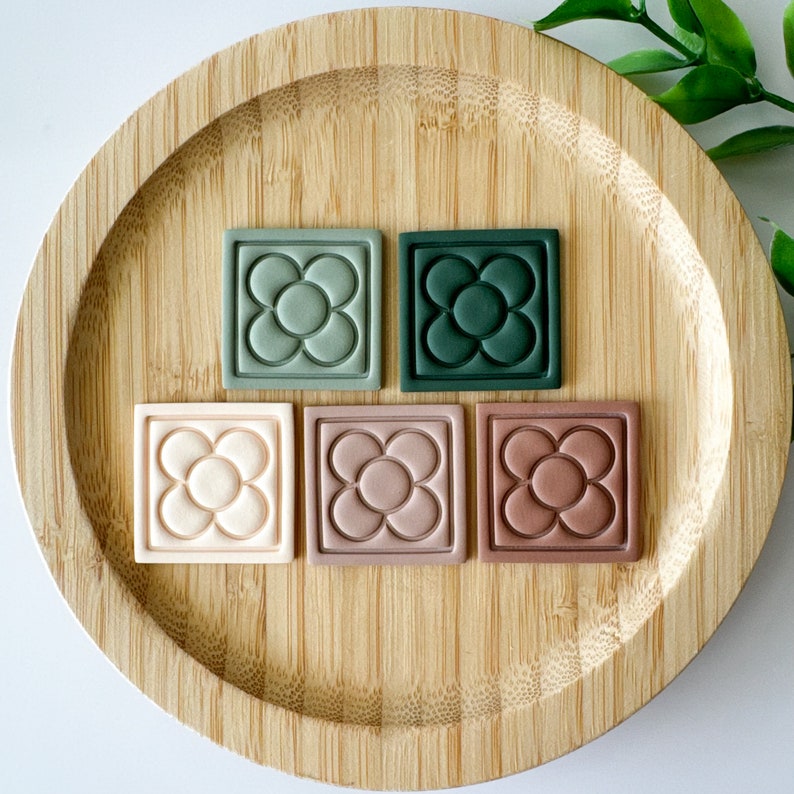 May include: Five square tiles featuring a raised five-petal flower design arranged in a row. The tiles are colored in shades of sage green, dark forest green, cream, dusty rose, and terracotta, each with a smooth, matte finish and border.
