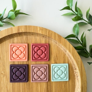 May include: Five ceramic tiles with a flower design in different colours. The tiles are arranged in a row on a wooden surface. The colours of the tiles are red, pink, purple, blue, and orange.