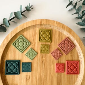 Può includere: Una collezione di formine per biscotti quadrate e a forma di diamante in varie dimensioni e colori, tra cui verde, oliva e rosso. Ogni formina presenta un design floreale. Le formine sono disposte su un vassoio di legno.