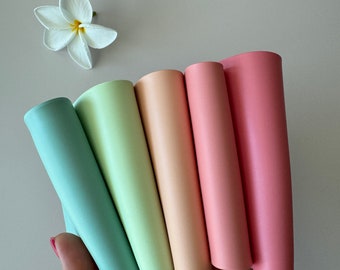 May include: Five smooth, cylindrical rolls of material in a gradient of pastel colors, including teal, light green, peach, light pink, and rose pink, arranged in a fan shape next to a single white five-petaled flower with a yellow center.