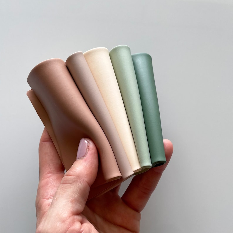May include: A set of five flexible, smooth, matte-finish silicone strips arranged in a fan shape. The colors transition from a warm terracotta brown to a light beige, followed by a soft sage green and a deeper forest green shade.