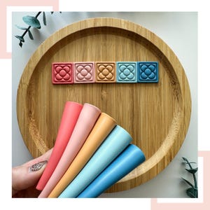 Puede incluir: Cinco mordedores de silicona en un arcoíris de colores: rosa, coral, melocotón, azul claro y azul. Los mordedores tienen forma de cono y están dispuestos en forma de abanico. Los mordedores están en una bandeja de madera con cinco bloques cuadrados de silicona con un diseño de flor en el centro. Los bloques están en el mismo orden de colores que los mordedores.