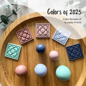 Peut inclure: Un plateau en bois avec sept boules d'argile de différentes couleurs : rose, bleu, vert, bleu clair, bleu foncé et deux nuances de rose. Les boules sont disposées en cercle autour d'une petite tuile carrée avec un motif de fleur. La tuile est également de couleur bleu clair. Le plateau est étiqueté « Colors of 2025 » et « Color Recipes of Sculpey Premo ».
