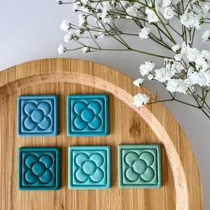 Puede incluir: Cuatro azulejos de cerámica azul verdoso con un diseño de flor estilizado. Los azulejos están dispuestos en dos filas de dos sobre una superficie de madera.